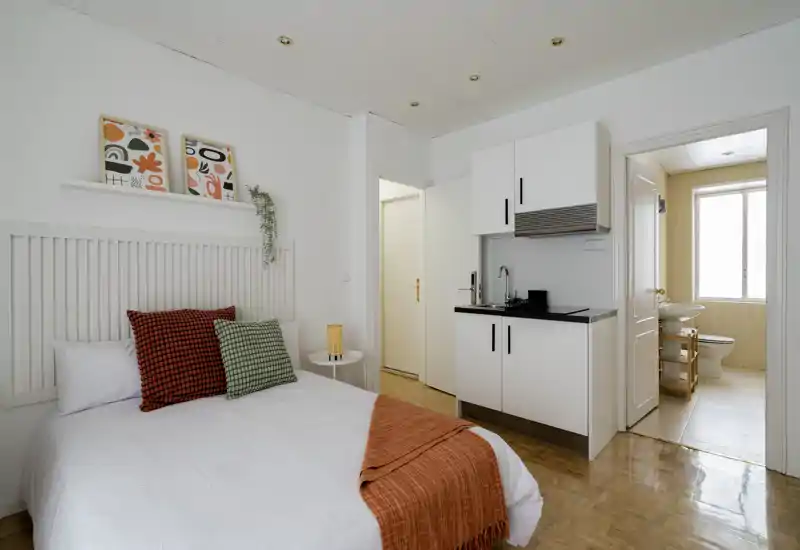 Bright, modern bedroom with a compact kitchenette. The photo shows a neatly made double bed with decorative pillows and a warm throw, plus a small sink and cabinets — ideal for a studio rental.