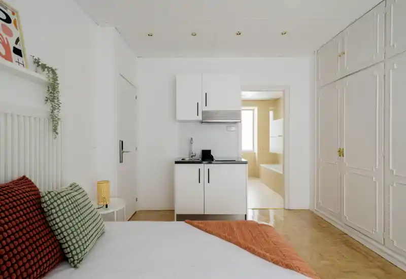 Bright, neatly styled studio bedroom with a small kitchenette area, large built-in wardrobe and a white, airy feel. Colorful cushions and an orange throw add warmth.