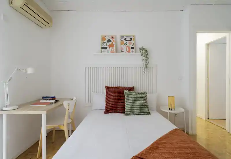 Bright, minimal bedroom with a neatly made double bed, colorful textured pillows and a small desk by the wall — ideal for a calm stay or working from the room.