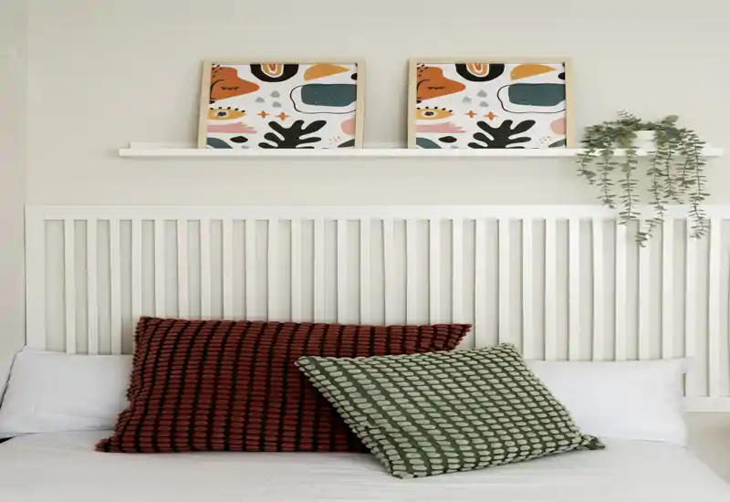 Primer plano de dormitorio luminoso con cabecero blanco de listones, cojines decorativos y cuadros minimalistas: espacio para dormir tranquilo y bien iluminado.