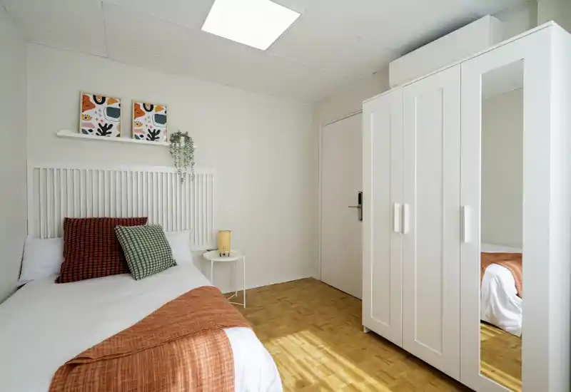 Dormitorio individual luminoso y ordenado con textiles cálidos, cojines decorativos y un gran armario blanco con espejo — ideal para un alquiler acogedor.