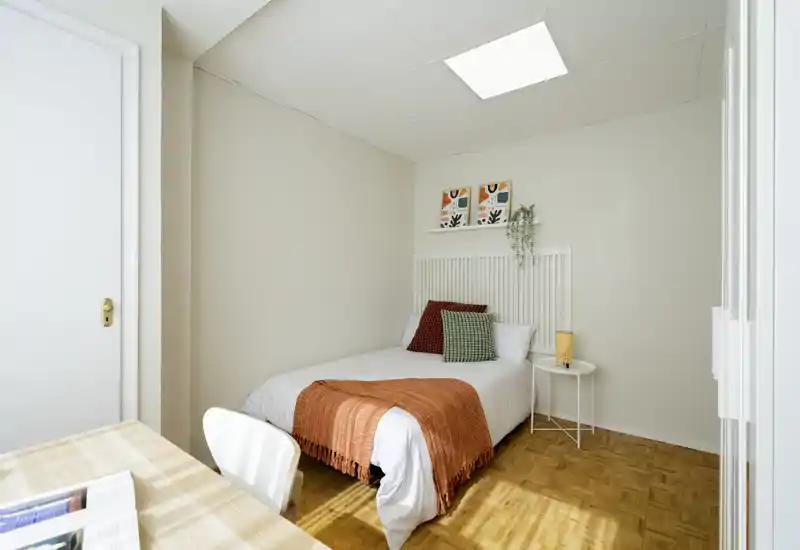 Dormitorio individual luminoso y bien presentado con la cama hecha, manta naranja y cojines decorativos. Una mesita auxiliar y una repisa en la pared completan el ambiente acogedor y minimalista, perfecto para un anuncio de alquiler.