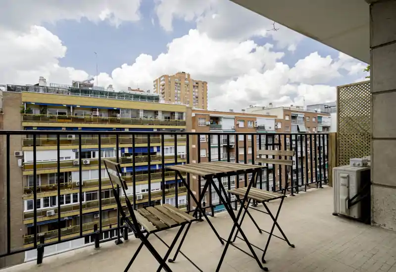 Sunny balcony with a small wooden bistro table and two chairs overlooking neighboring apartment buildings — ideal for morning coffee or relaxing outdoors.