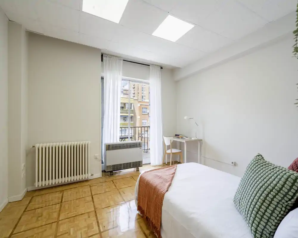 Bright, minimal bedroom with a comfortable single/double bed dressed in white linens and a coral throw, a small desk by the balcony window, and parquet flooring providing warm contrast.
