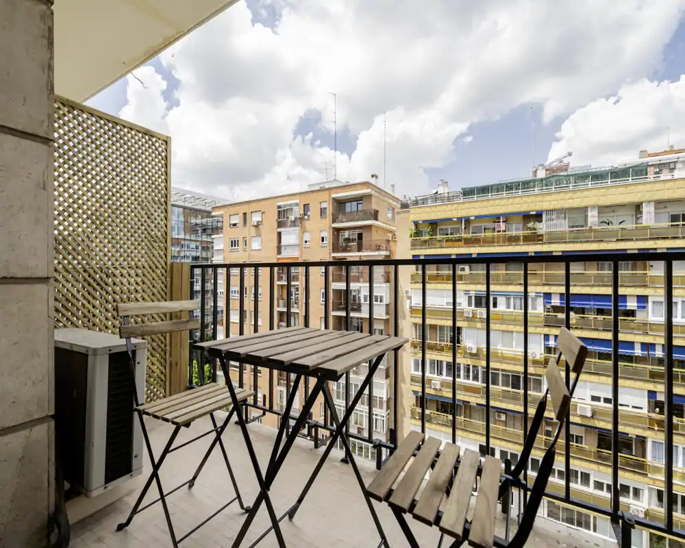 Sunny urban balcony with a small wooden bistro table and folding chairs, ideal for morning coffee and city views. Bright daylight and open railings create an inviting outdoor spot.