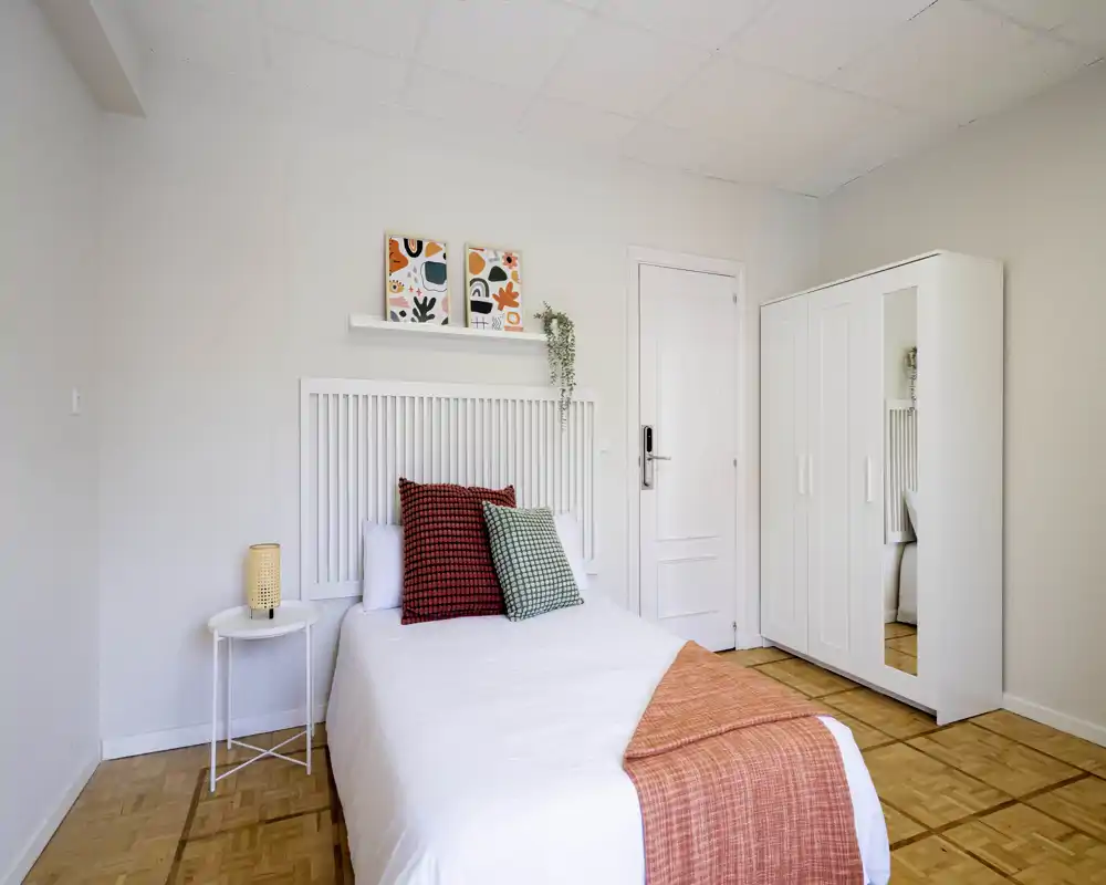 Bright, minimal bedroom with a neatly made double bed, colorful cushions and a matching throw; white wardrobe with mirrored door and simple bedside table complete the cozy setup.
