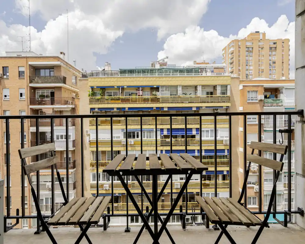 Sunny urban balcony with a small wooden bistro table and two chairs, overlooking neighboring apartment buildings — ideal for morning coffee or a quiet city break.