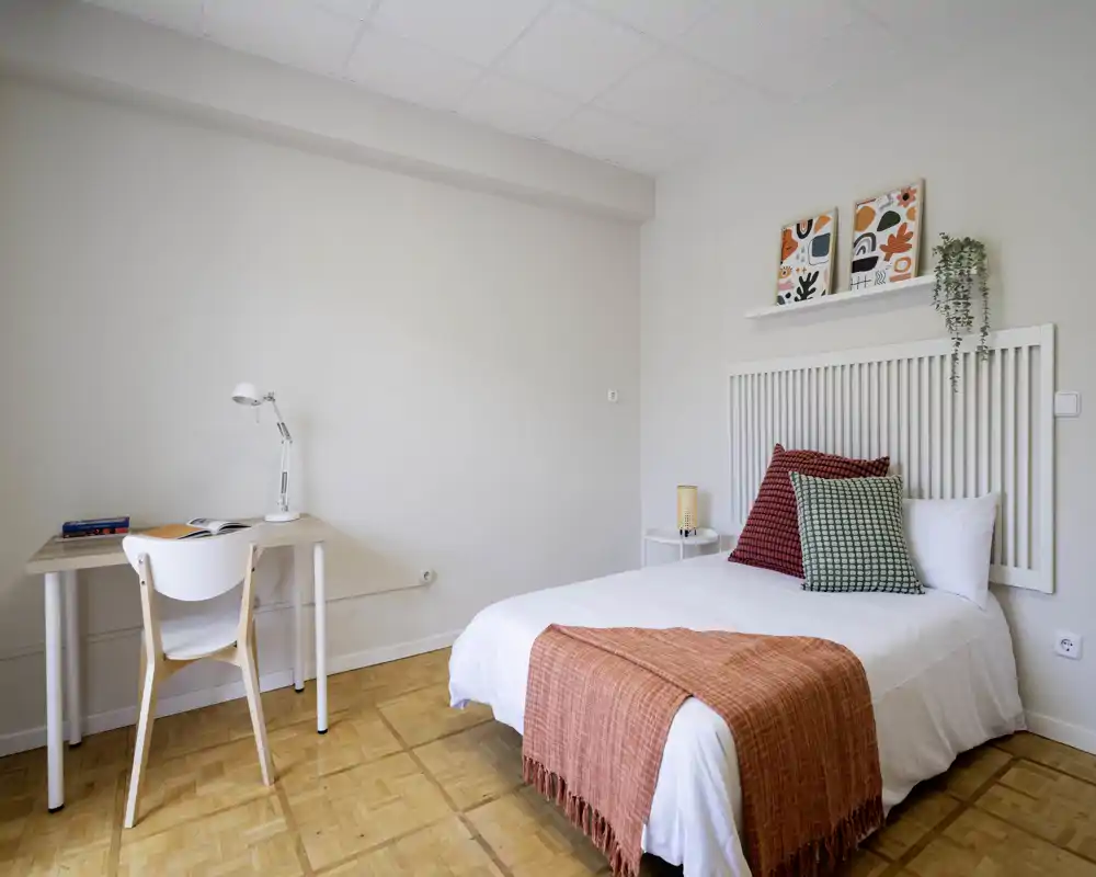 Bright, minimal bedroom with a neatly made single/double bed dressed in white linens, colorful throw and cushions, and a small desk by the wall — ideal for a calm stay or student flat.