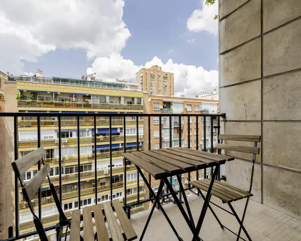 Sunny city balcony with a compact wooden bistro table and two folding chairs, overlooking neighboring apartment buildings — ideal for morning coffee or an evening drink.