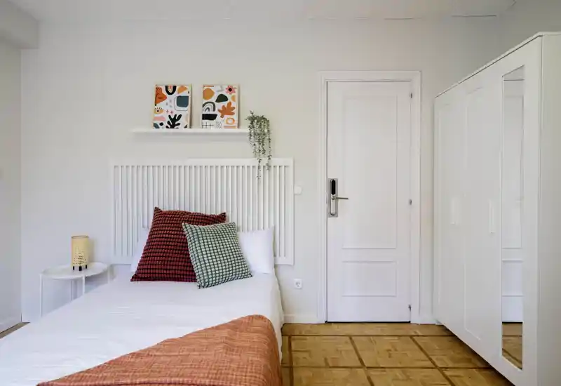 Bright, neatly styled single bedroom with a white bed, colorful cushions and a matching orange throw. The room features a white wardrobe with mirrored door, minimalist bedside table and simple wall decor for a fresh, modern look.