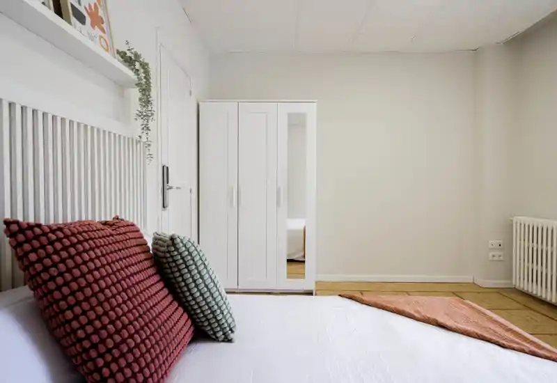 Bright, minimal bedroom with textured cushions, white wardrobe and a cozy throw on the bed — calm neutral palette and tidy styling.