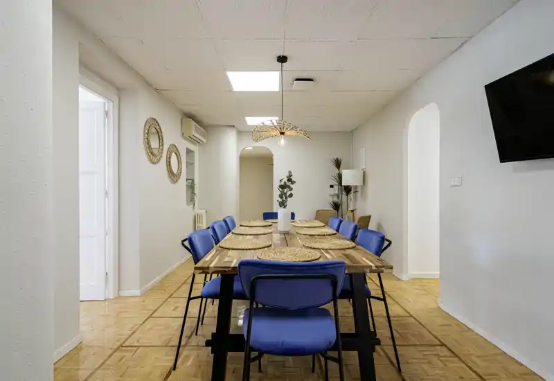 Comedor luminoso y decorado con una larga mesa de madera, sillas azules y salvamanteles trenzados bajo una lámpara colgante decorativa — ideal para comidas en grupo.