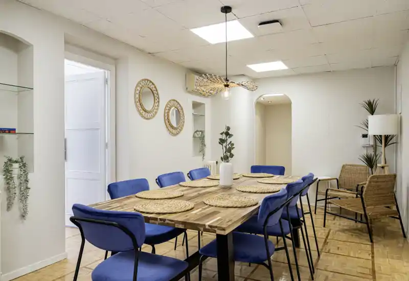 Comedor luminoso y estilizado con una gran mesa de madera para ocho, sillas tapizadas en azul y detalles naturales trenzados que crean un espacio moderno y acogedor.