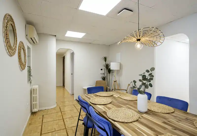 Área de comedor luminosa y cuidada con una gran mesa de madera, seis sillas azules, manteles individuales trenzados y una lámpara colgante decorativa — perfecta para comer en grupo.