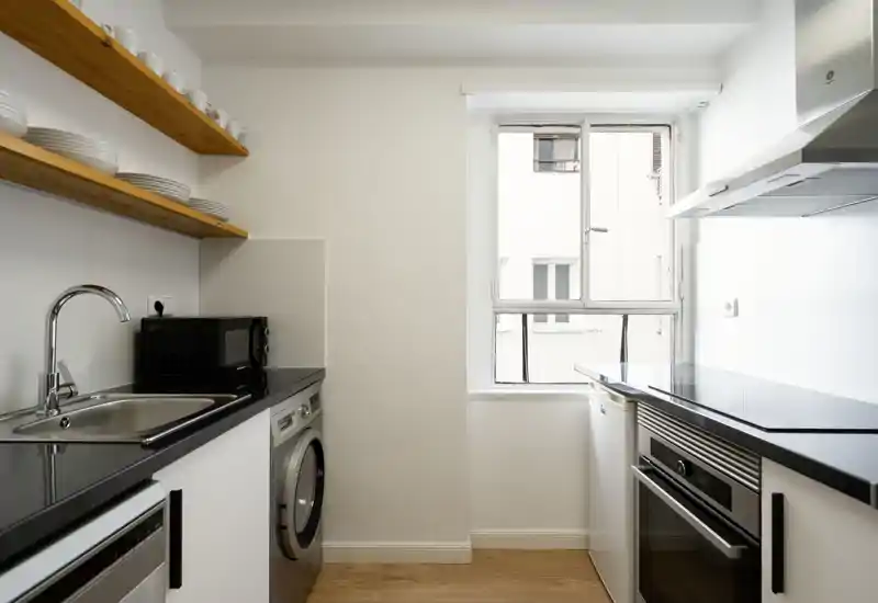 Cocina moderna y compacta con encimeras negras, horno integrado, placa de inducción y fregadero bajo estantes de madera; ventana luminosa aporta luz natural.