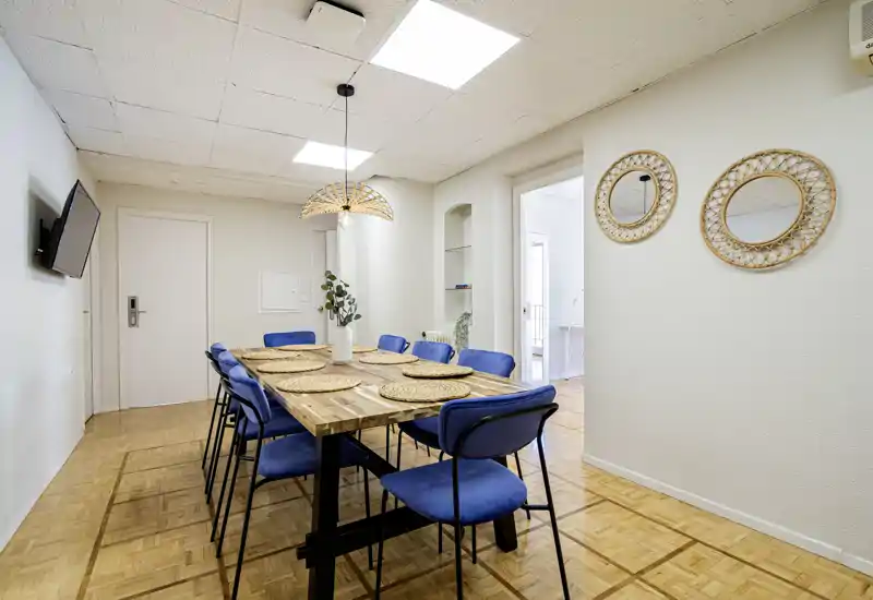 Comedor luminoso y cuidado con mesa de madera para ocho, sillas tapizadas azules, salvamanteles trenzados y lámpara colgante decorativa — ideal para comidas en grupo.