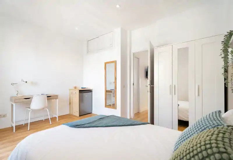Bright, minimal bedroom with a well-made bed in the foreground, wooden flooring, built-in wardrobe and a small desk—ideal for a comfortable stay.