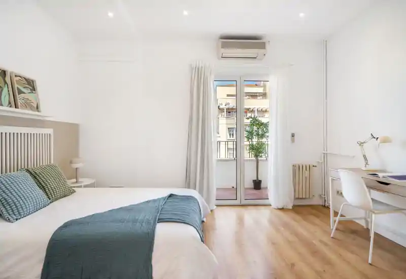 Bright, modern bedroom with a double bed dressed in white linens and blue accents, direct access to a small balcony and a compact desk area — ideal for comfortable short or long stays.