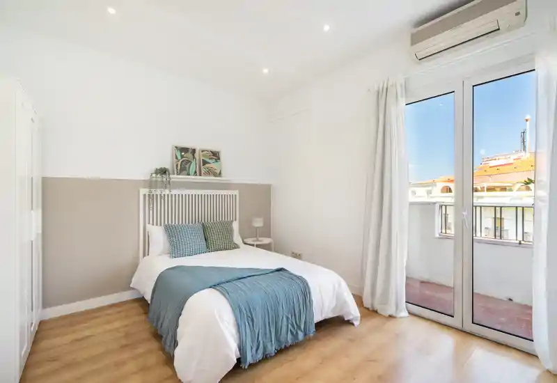 Bright, modern bedroom with a double bed, light wood floor and large glass doors leading to a small balcony. Soft textiles and cushions create a comfortable, airy atmosphere ideal for rental listings.