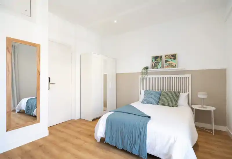 Bright, minimalist bedroom with a comfortable double bed dressed in white linens, a blue throw and patterned cushions; wardrobe, bedside table and a full-length mirror complete the simple, airy layout.