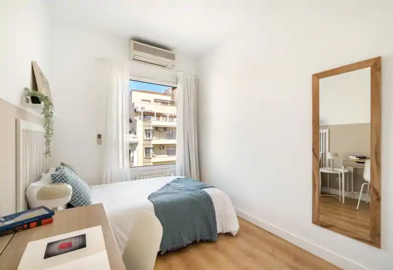 Chambre lumineuse et minimaliste avec lit double, textiles doux et miroir en pied. La lumière naturelle provenant de la grande fenêtre met en valeur le sol en bois et la décoration neutre.