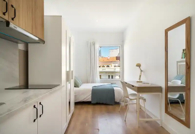 Bright, neatly styled studio bedroom with a single bed, workspace and a small kitchenette area; wood floors and neutral decor create a calm, modern atmosphere.