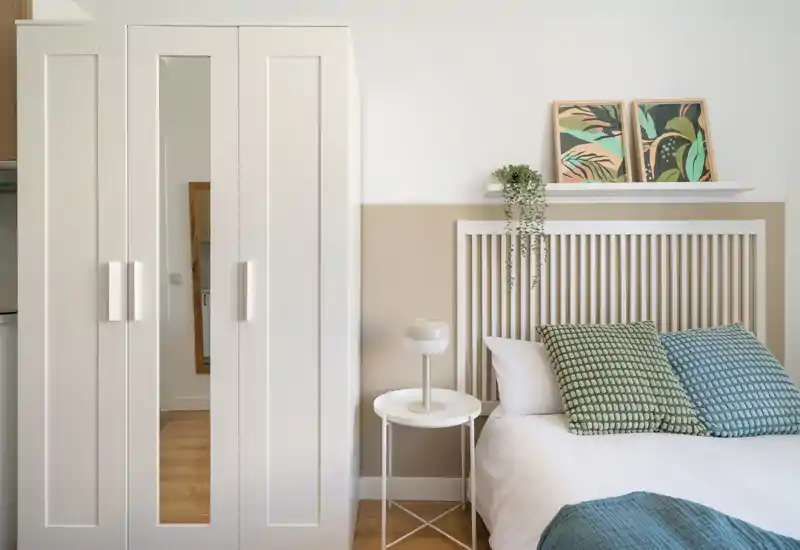 Bright, modern bedroom with a white slatted headboard, patterned cushions and a compact bedside table. The neutral palette and simple decor create a calm, inviting sleeping area.