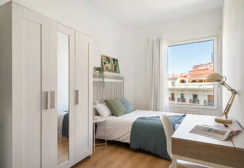 Bright, well-styled single bedroom with a comfortable double bed, decorative cushions and a desk by the window overlooking the city — ideal for a calm, sunny stay.