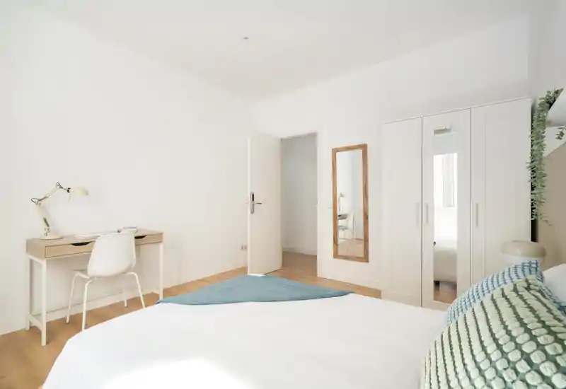 Dormitorio luminoso y minimalista con una cama grande en primer plano, armario blanco, escritorio y suelo de madera clara. La habitación resulta fresca y acogedora, con textiles suaves y líneas limpias.