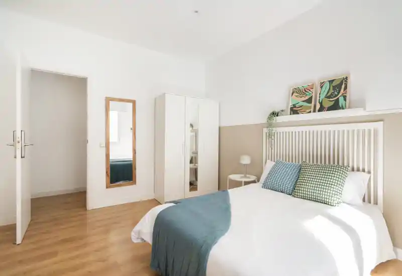 Dormitorio luminoso y aireado con cabecero de listones blanco, cama doble bien hecha, cojines decorativos y una manta azul. Suelo de madera clara y armario sencillo para un aspecto moderno y pulcro, perfecto para anuncios.