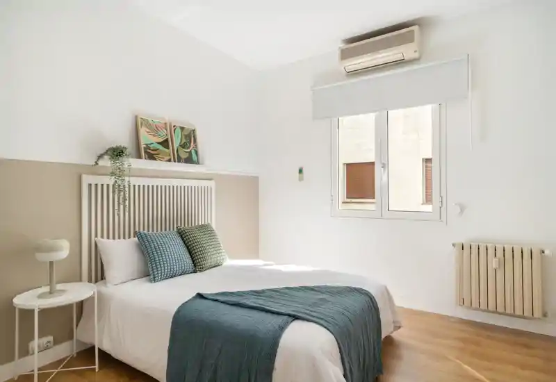 Dormitorio luminoso y minimalista con cama doble en ropa blanca, cojines decorativos y manta verde azulado, luz natural desde la ventana y plantas como detalle.