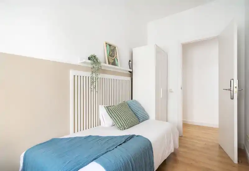 Dormitorio individual luminoso y minimalista con cama bien hecha, cojines decorativos y un pequeño armario — aireado, bien iluminado y cuidado.