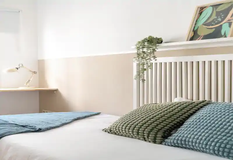 Dormitorio luminoso y minimalista con cojines texturizados y cama bien hecha bajo una cabecera de lamas; la luz natural y los tonos neutros crean un ambiente sereno.