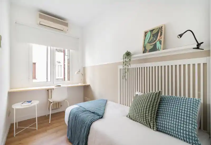 Dormitorio individual luminoso y bien decorado con cama cómoda, cojines texturizados y plaid, y pequeño escritorio junto a la ventana — ideal para estudiante o una persona sola.