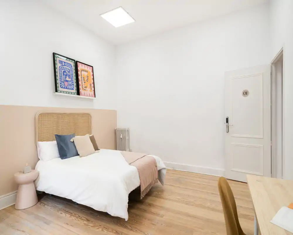 Chambre lumineuse et minimaliste avec tête de lit en osier, linge de lit doux et coussins décoratifs. Parquet clair et accents pastel neutres pour une ambiance calme et aérée idéale pour la location.