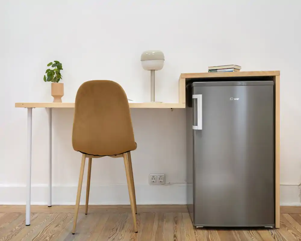 Coin kitchenette compact avec un petit réfrigérateur en acier inox sous un bureau en bois, une chaise simple, une lampe et une plante — lumineux, minimal et fonctionnel.