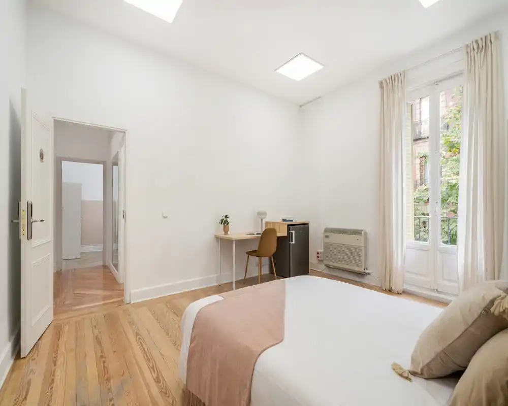 Chambre lumineuse et minimaliste avec un lit double confortable, une couverture douce et des coussins, un petit bureau et une chaise près d'une grande fenêtre offrant une belle lumière naturelle.