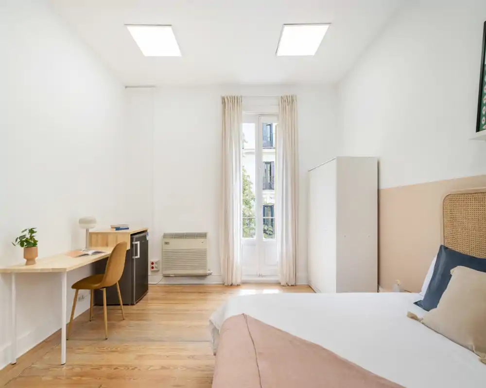 Chambre lumineuse et minimaliste avec un lit bien fait, lumière naturelle par une grande fenêtre, petit bureau et armoire — idéale pour un séjour confortable.