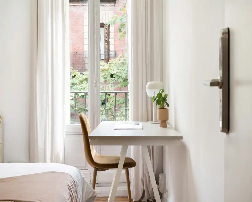 Coin chambre lumineux et minimaliste avec un bureau blanc devant une grande fenêtre, une chaise moutarde, une lampe et une petite plante. Le lit est partiellement visible à gauche, donnant une ambiance calme et aérée, idéale pour une location urbaine compacte.