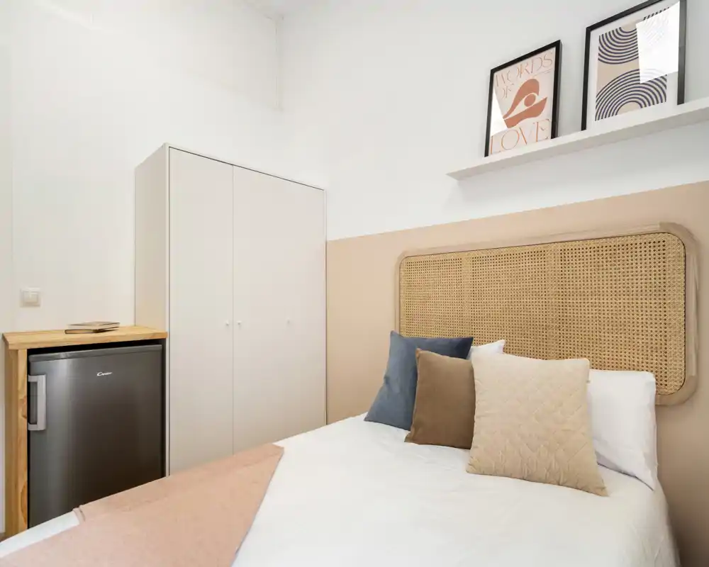 Chambre lumineuse et soigneusement mise en scène avec une tête de lit tressée et des coussins décoratifs ; armoire compacte et petit réfrigérateur visibles, idéale pour un séjour cosy.