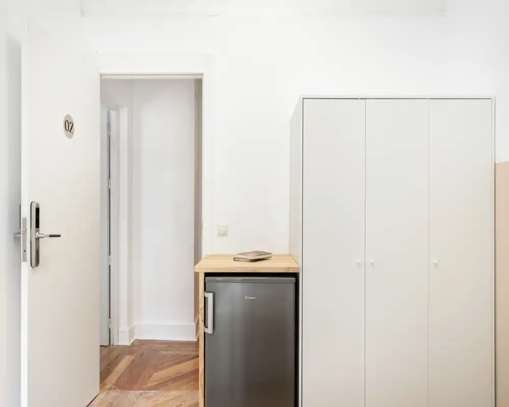Entrée lumineuse et minimaliste avec une armoire blanche, un petit mini-frigo inox sur un plan en bois et une porte ouverte vers un couloir intérieur — espace d'entrée propre et fonctionnel.