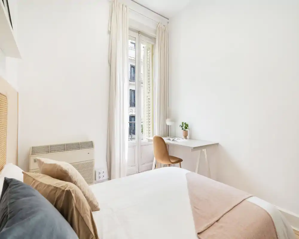 Chambre lumineuse et minimaliste avec un lit double bien fait, textiles neutres et un petit bureau près de la fenêtre laissant entrer la lumière naturelle.