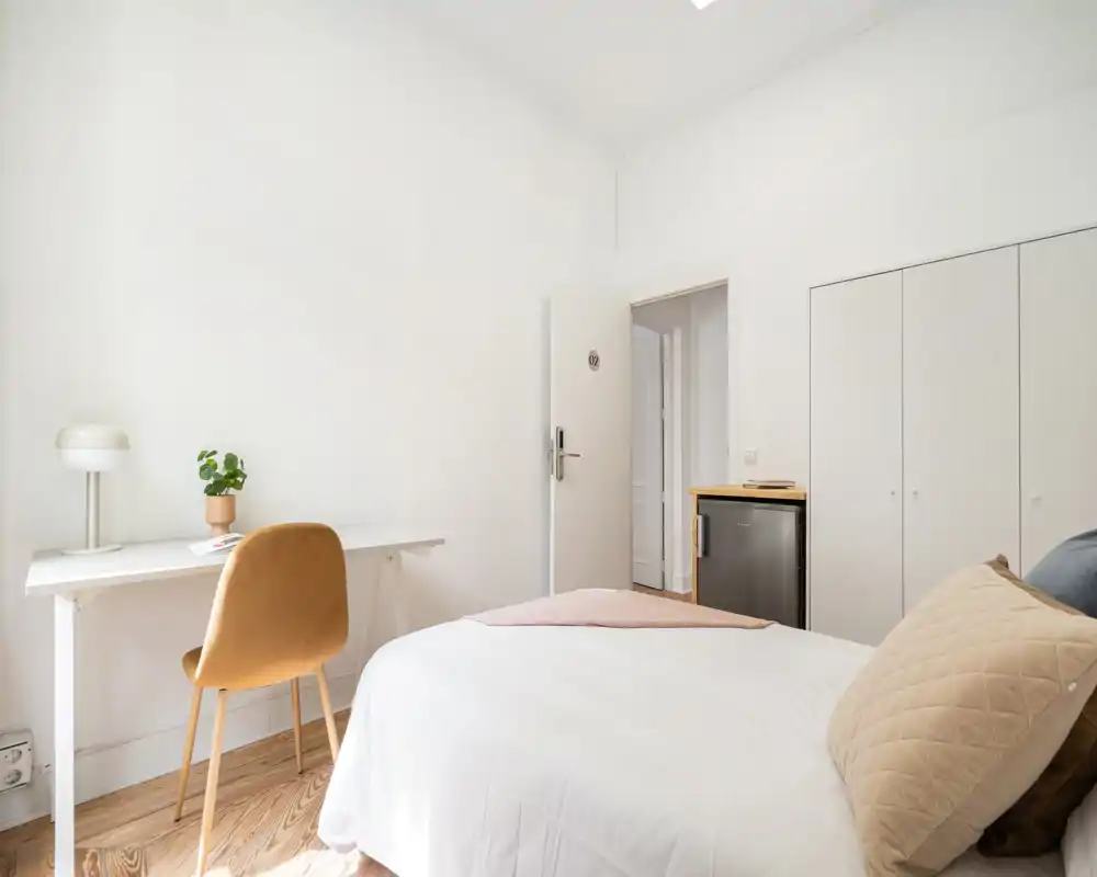 Chambre lumineuse et minimaliste avec lit bien fait, textiles doux et un petit espace de travail près du mur—parfaite pour de courts séjours.