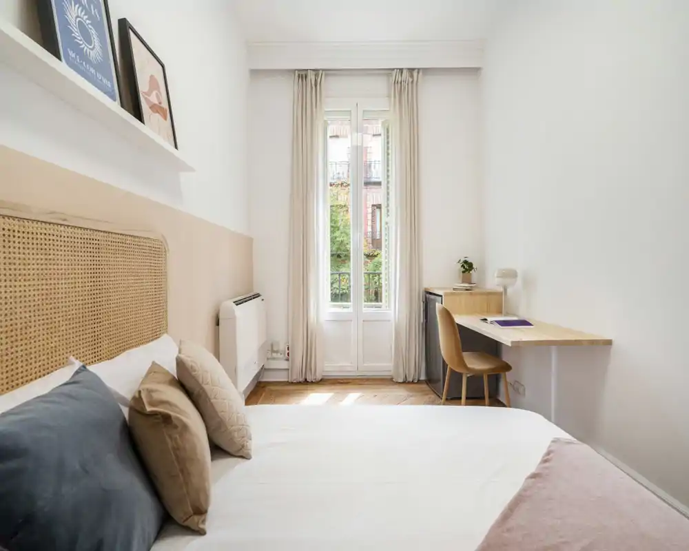 Chambre lumineuse et minimaliste avec tête de lit en rotin, coussins superposés et un petit bureau près de la fenêtre. La grande fenêtre inonde la pièce de lumière naturelle et la palette douce crée une ambiance reposante.