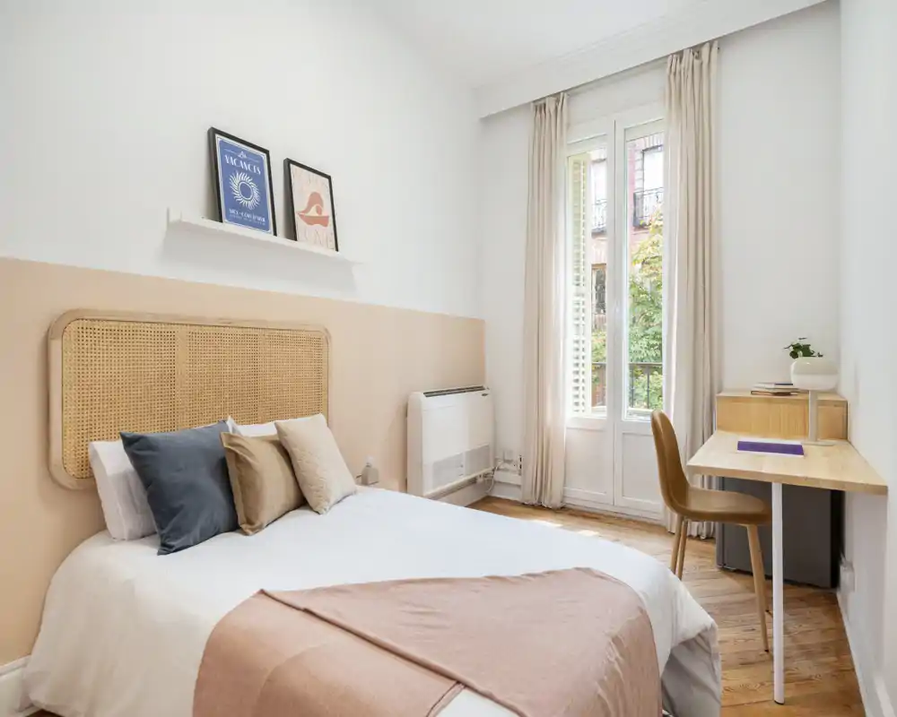 Chambre lumineuse et élégante avec tête de lit en cannage, coussins superposés et un coin bureau soigné près de la fenêtre — idéale pour se reposer ou travailler à distance.