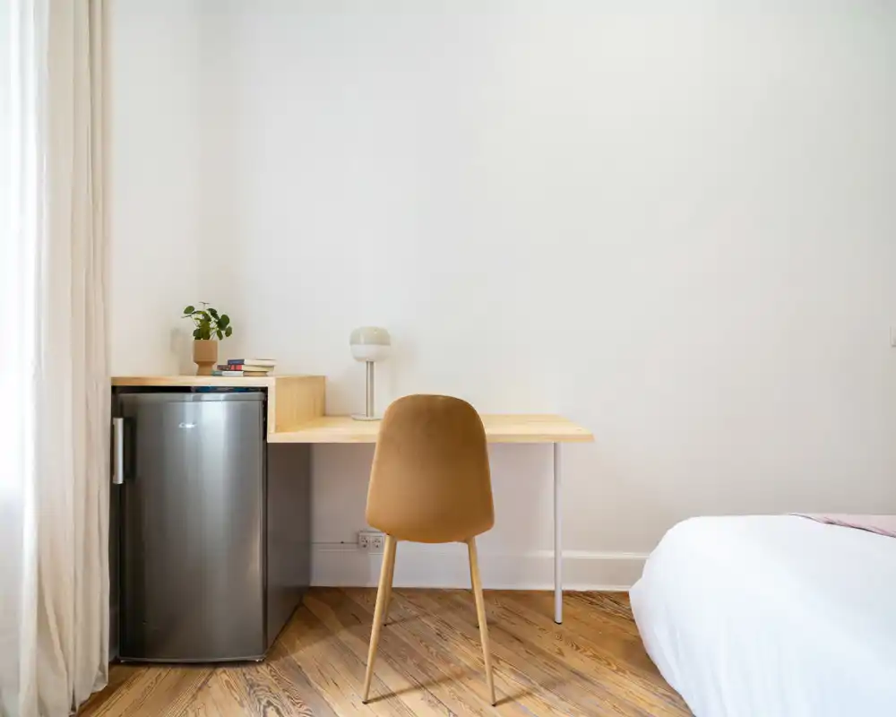 Coin de chambre lumineux et minimaliste avec le bord du matelas visible, bureau en bois et petit réfrigérateur — propre, moderne et fonctionnel pour de courts séjours.