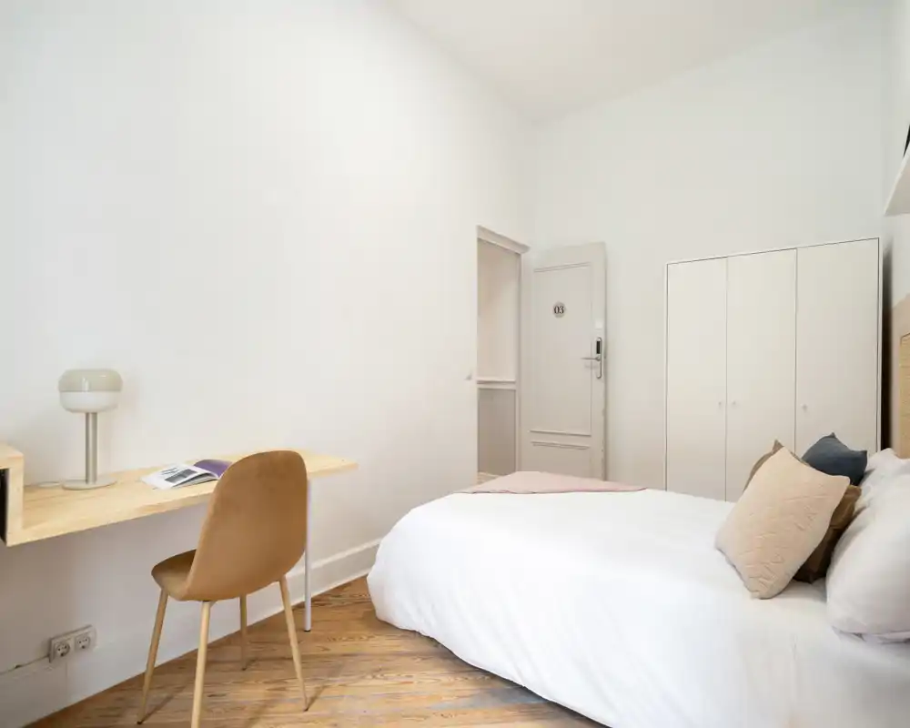 Chambre lumineuse et minimaliste avec un lit double confortable, linge de lit neutre et un petit bureau mural idéal pour travailler ou lire.