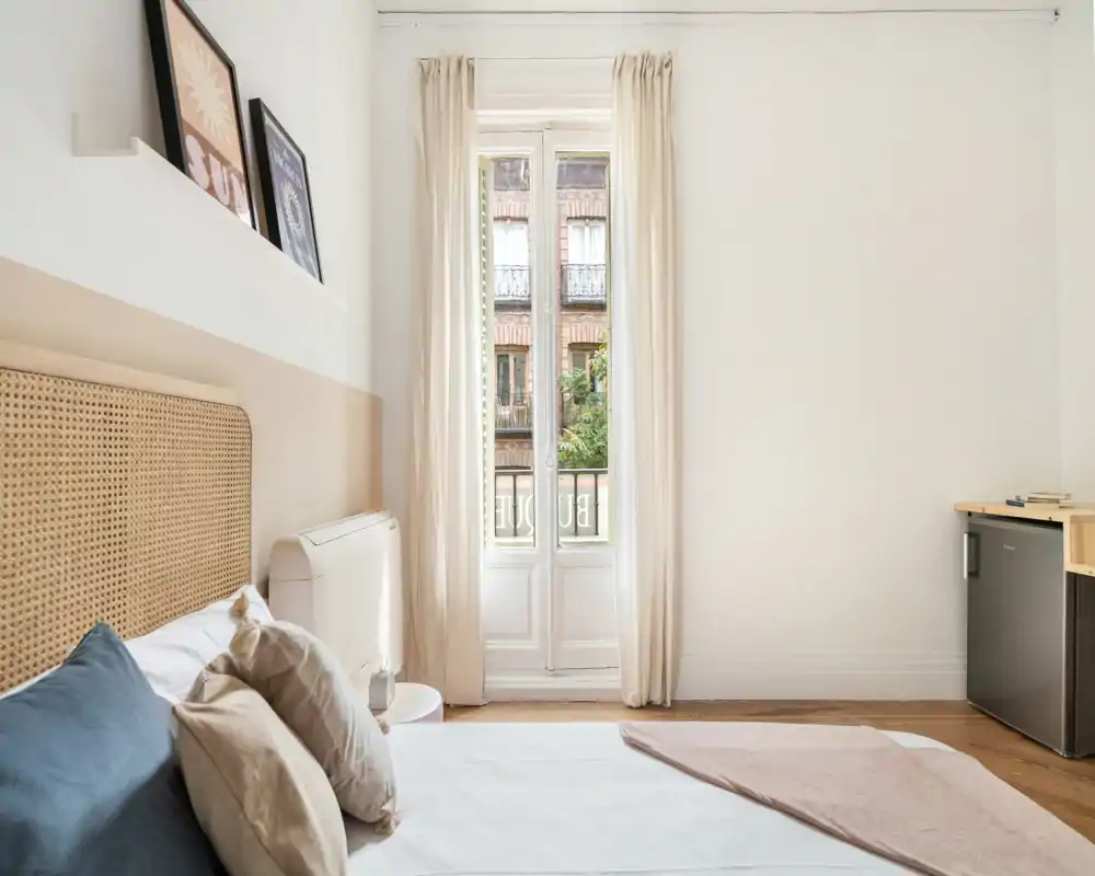 Chambre lumineuse et minimaliste avec tête de lit en rotin, linge doux et grande fenêtre laissant entrer une belle lumière naturelle — idéale pour des annonces locatives recherchant une ambiance calme et aérée.