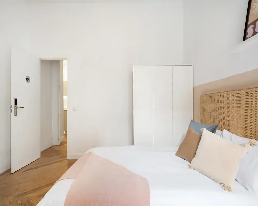 Chambre lumineuse et minimaliste avec tête de lit tressée, linge de lit blanc et accents pastel. Grand lit visible, armoire et sol en bois chaleureux, parfaite pour un séjour reposant.