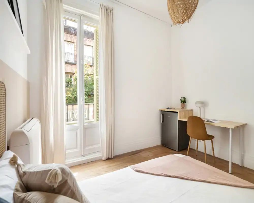 Chambre lumineuse et minimaliste avec un lit confortable, une grande fenêtre laissant entrer la lumière naturelle et un petit coin bureau — idéale pour se détendre ou travailler à distance.